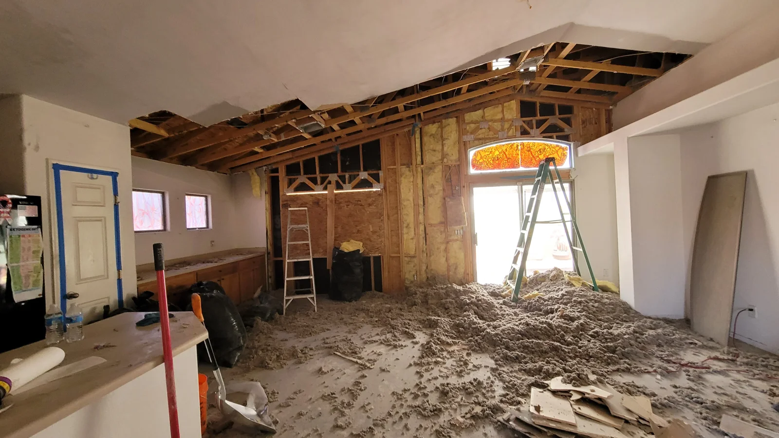 Gutted room with exposed wood roof rafters, loose insulation and drywall debris covering the floor, and debris bags