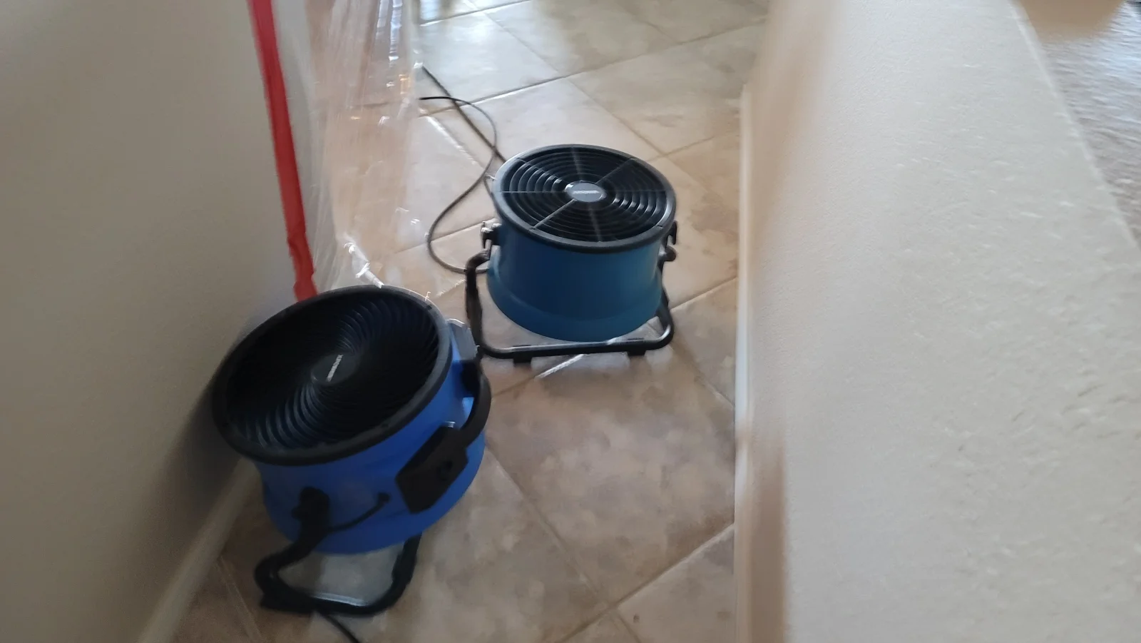 Two blue air movers drying a beige ceramic tile floor beside plastic sheeting taped to a wall
