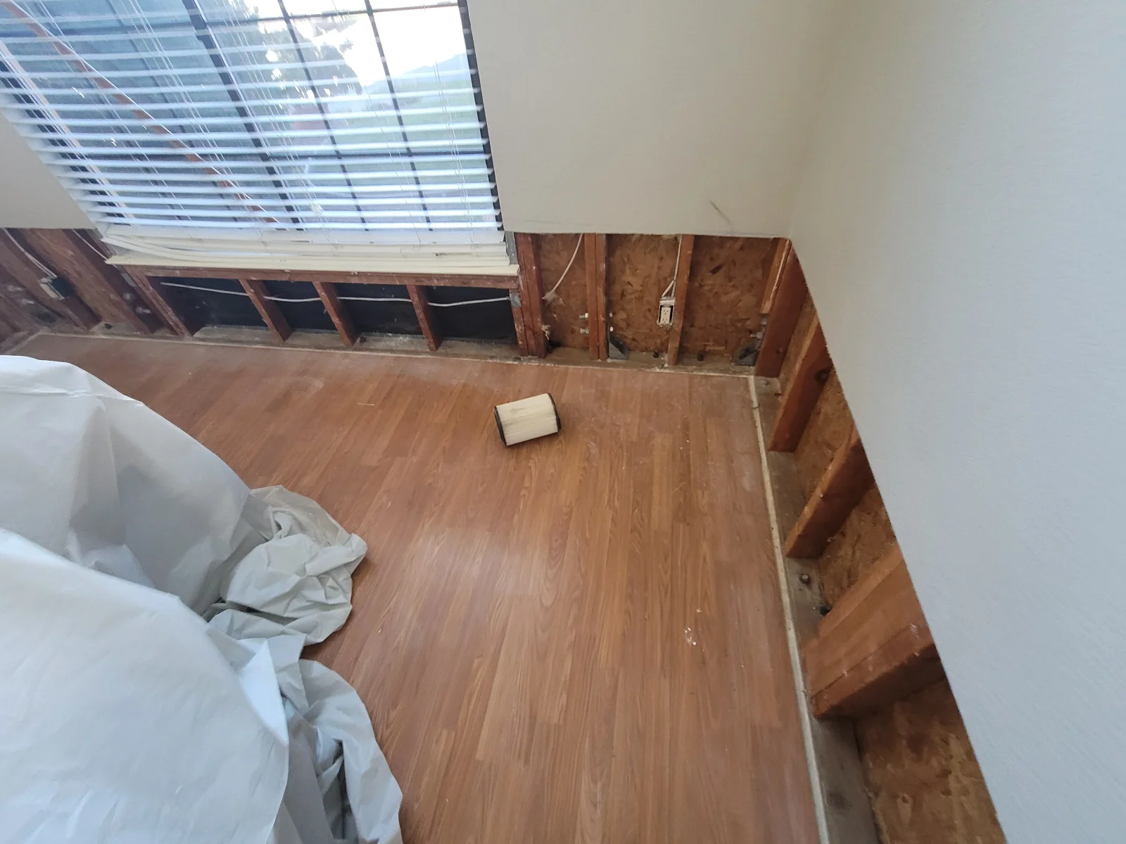 Room with drywall removed to expose wood studs, sheathing, and wiring, with laminate flooring and plastic sheeting