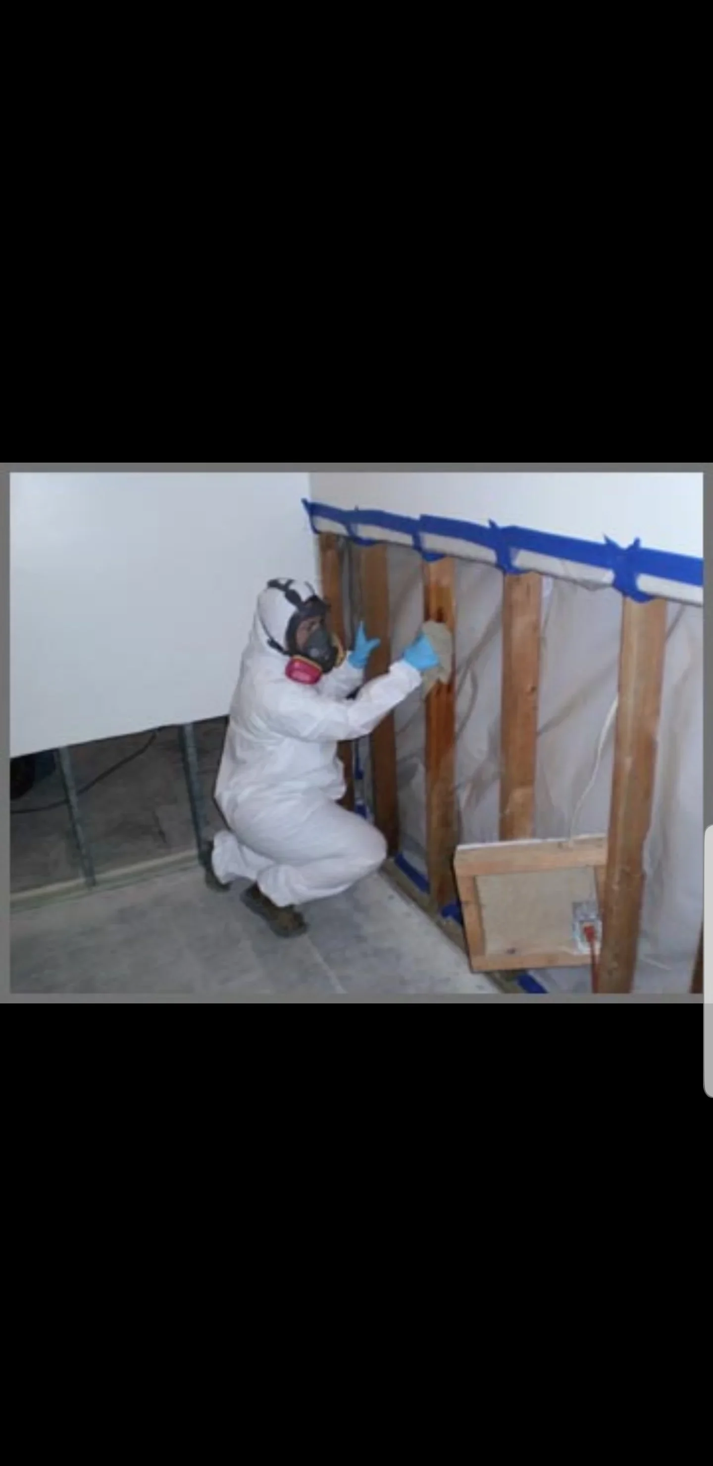Technician in protective suit and respirator cleaning exposed wood wall studs with plastic sheeting and blue tape containment