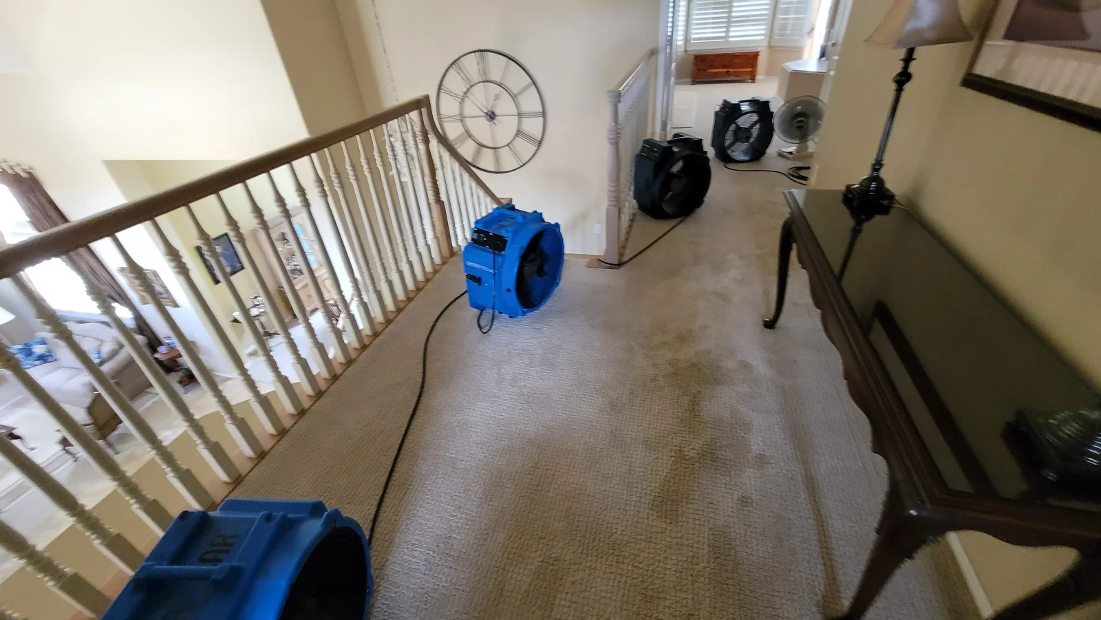 Blue and black air movers drying a beige carpeted upstairs hallway with water staining near a wood stair railing