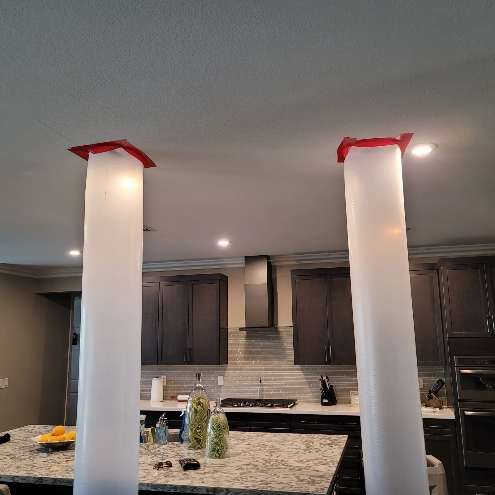 White plastic-wrapped columns sealed with red tape in a kitchen with dark cabinets and granite island