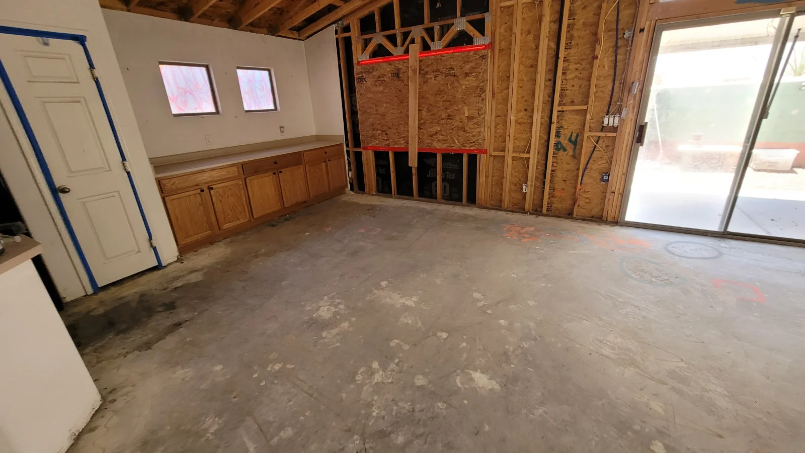 Gutted room with exposed wood wall framing, OSB sheathing, bare concrete floor, and oak cabinets along one wall