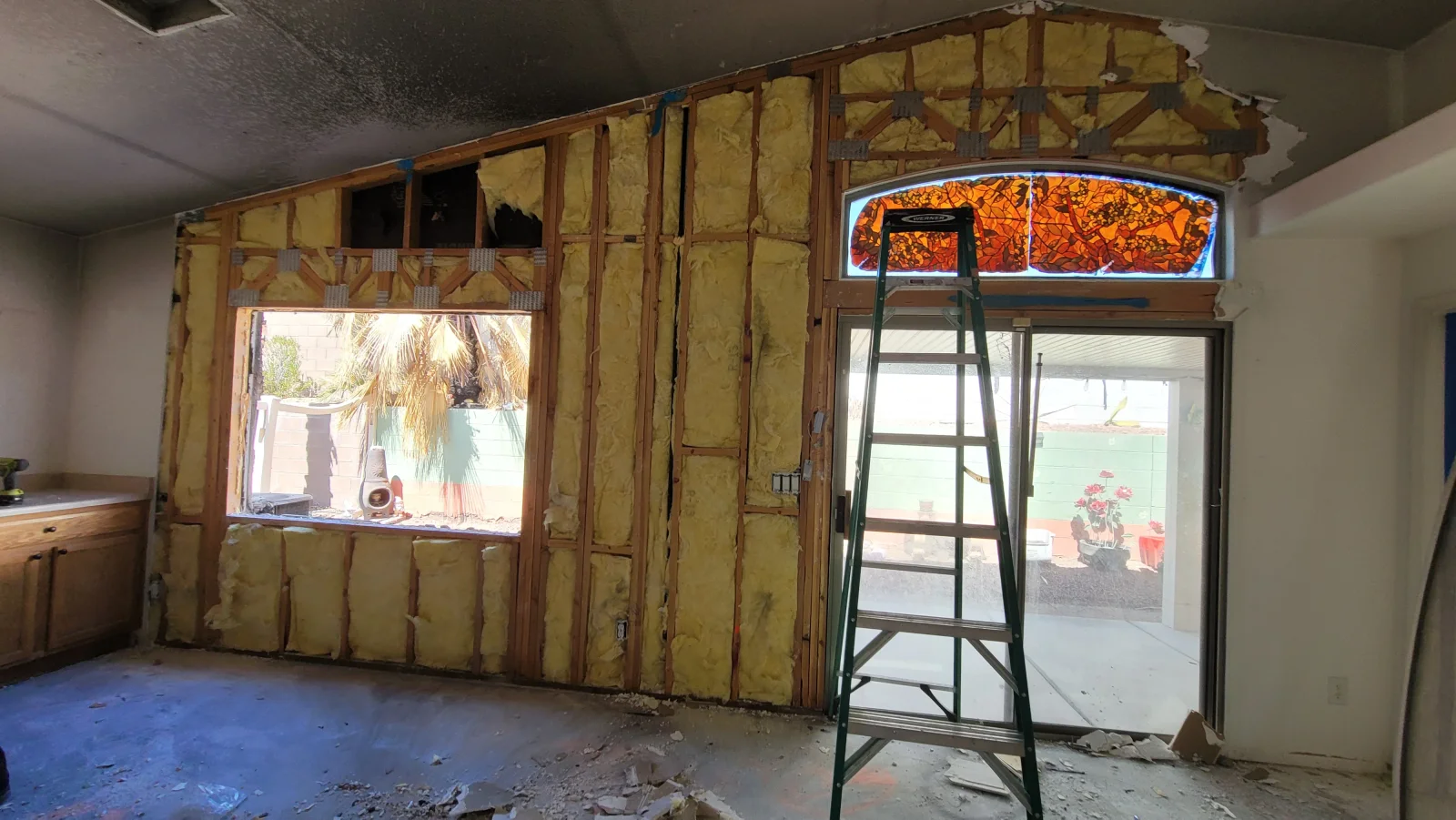 Open wall cavity with wood studs, yellow fiberglass insulation, and a stained glass transom above a sliding glass door