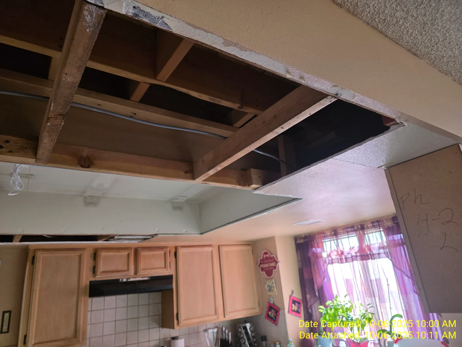 Open kitchen ceiling with removed drywall exposing wood joists, cross bracing, and electrical cable above oak cabinets