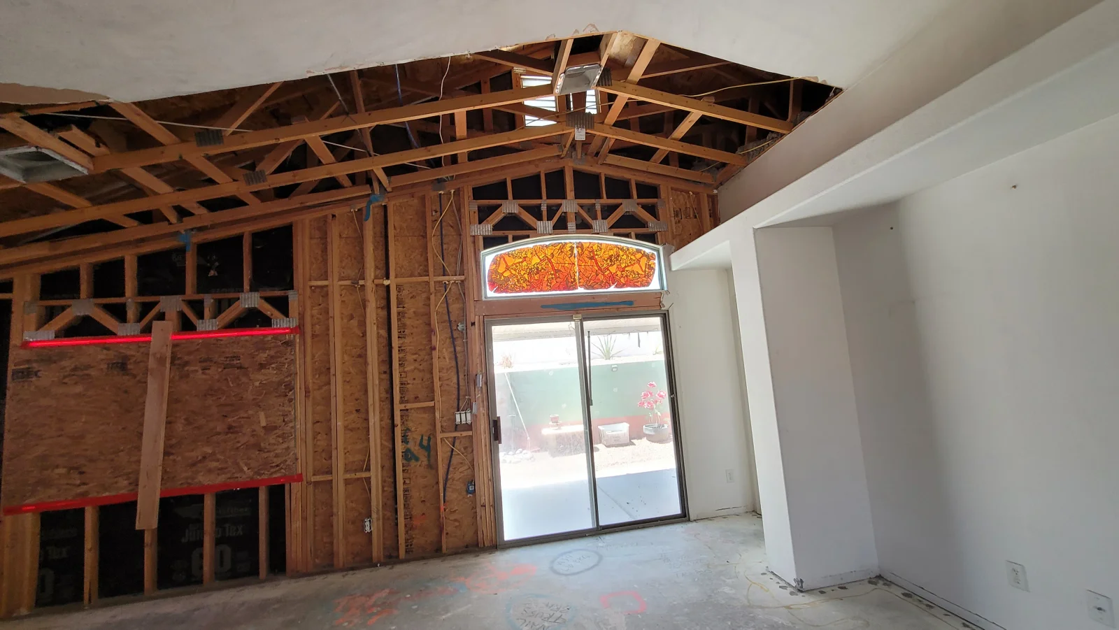 Gutted interior with exposed wood trusses, OSB-sheathed wall sections, wood studs, and an arched stained glass transom above a sliding glass door