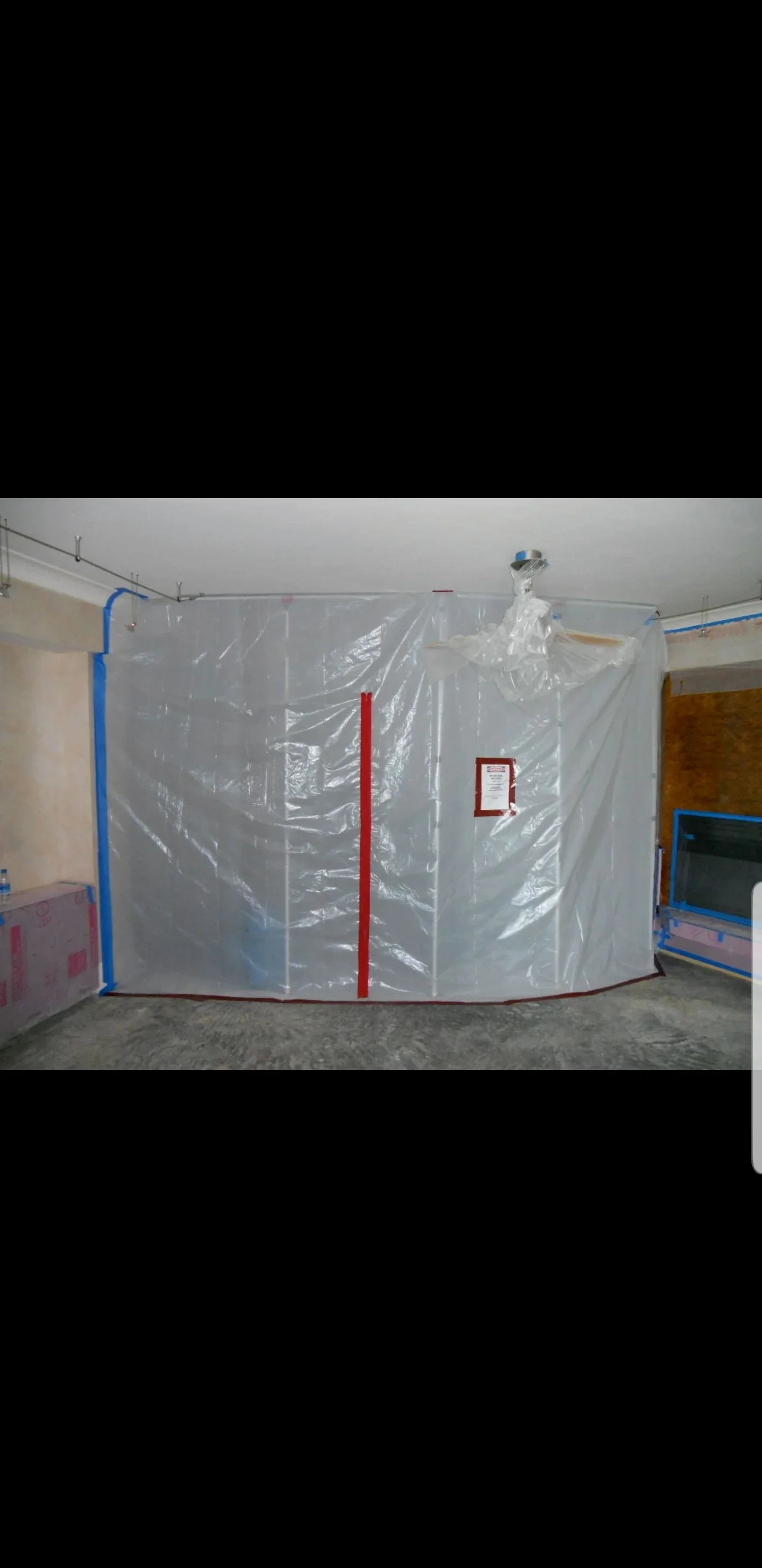 Clear plastic containment sheeting taped with blue painter's tape across an interior room, with a red seam tape and placard on the barrier