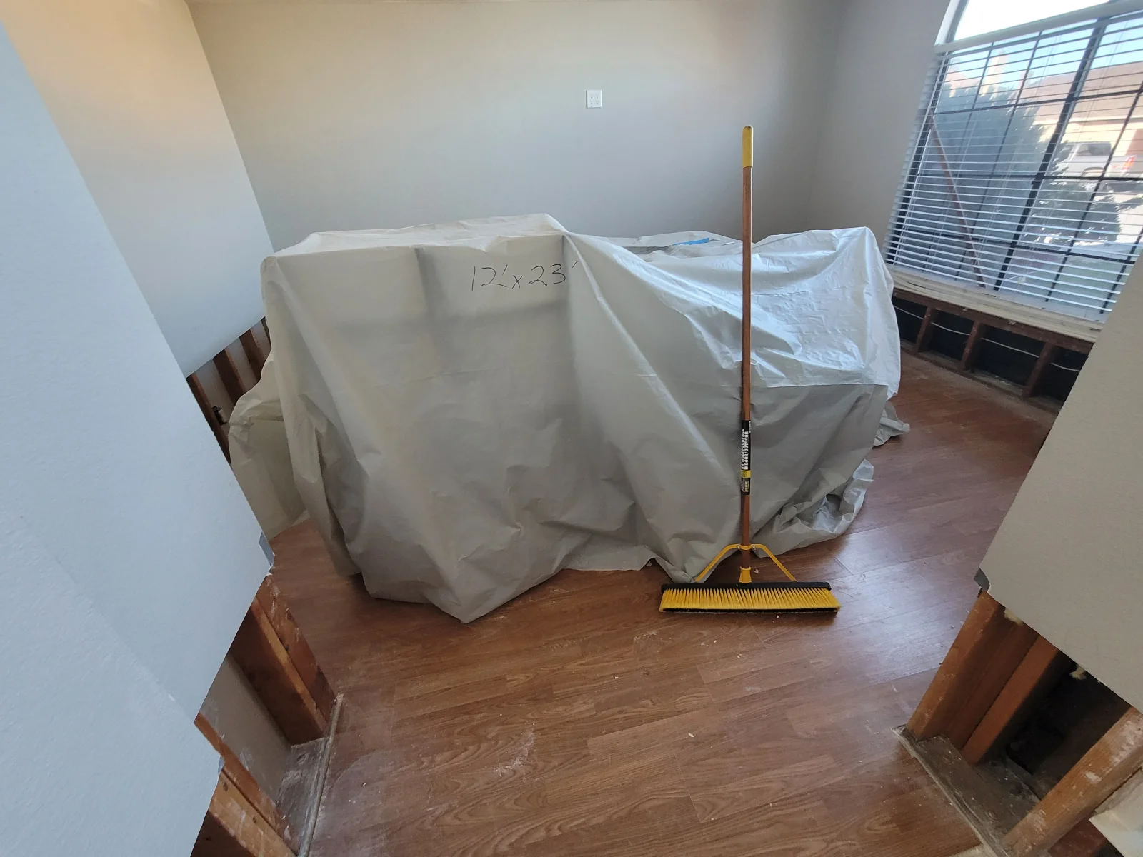 Room with laminate flooring removed to wood subfloor, contents covered in plastic sheeting, and yellow push broom on bare floor